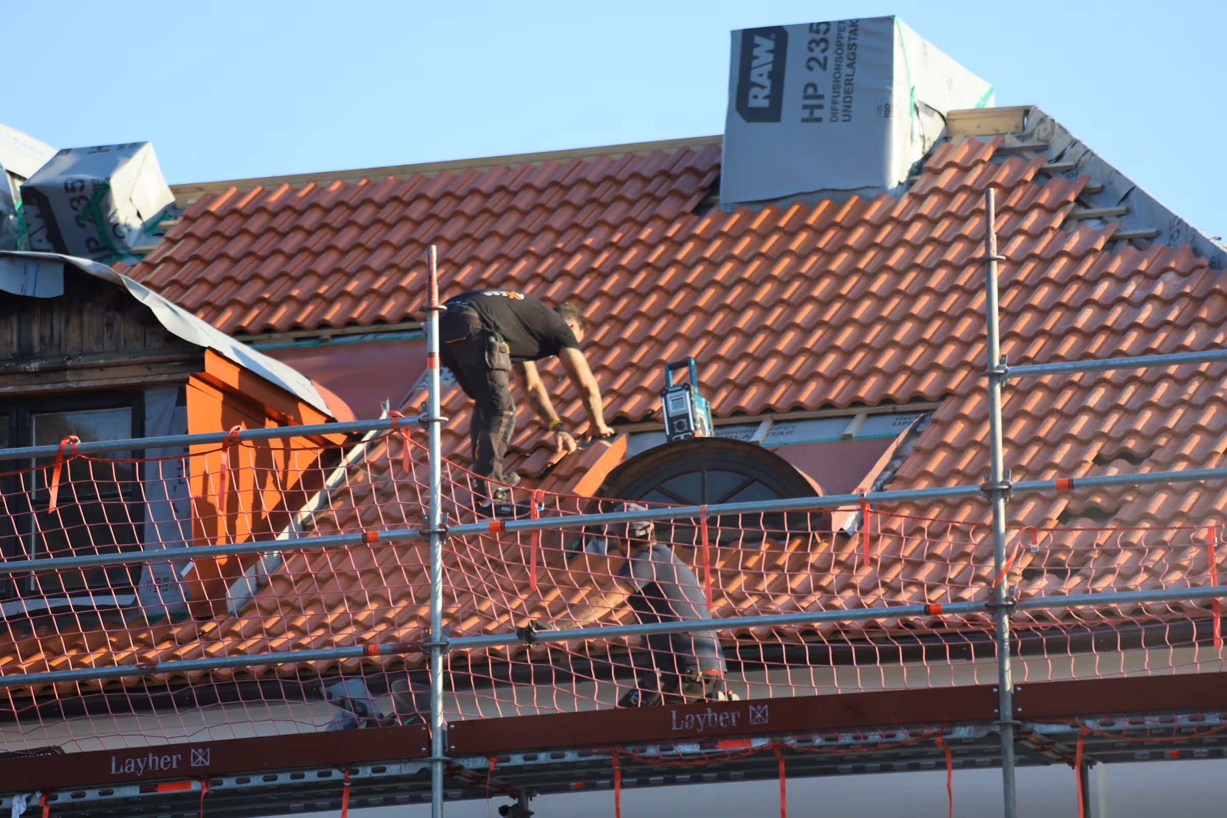 Worker repairing red tile roof on a house, standing on scaffolding with safety netting.