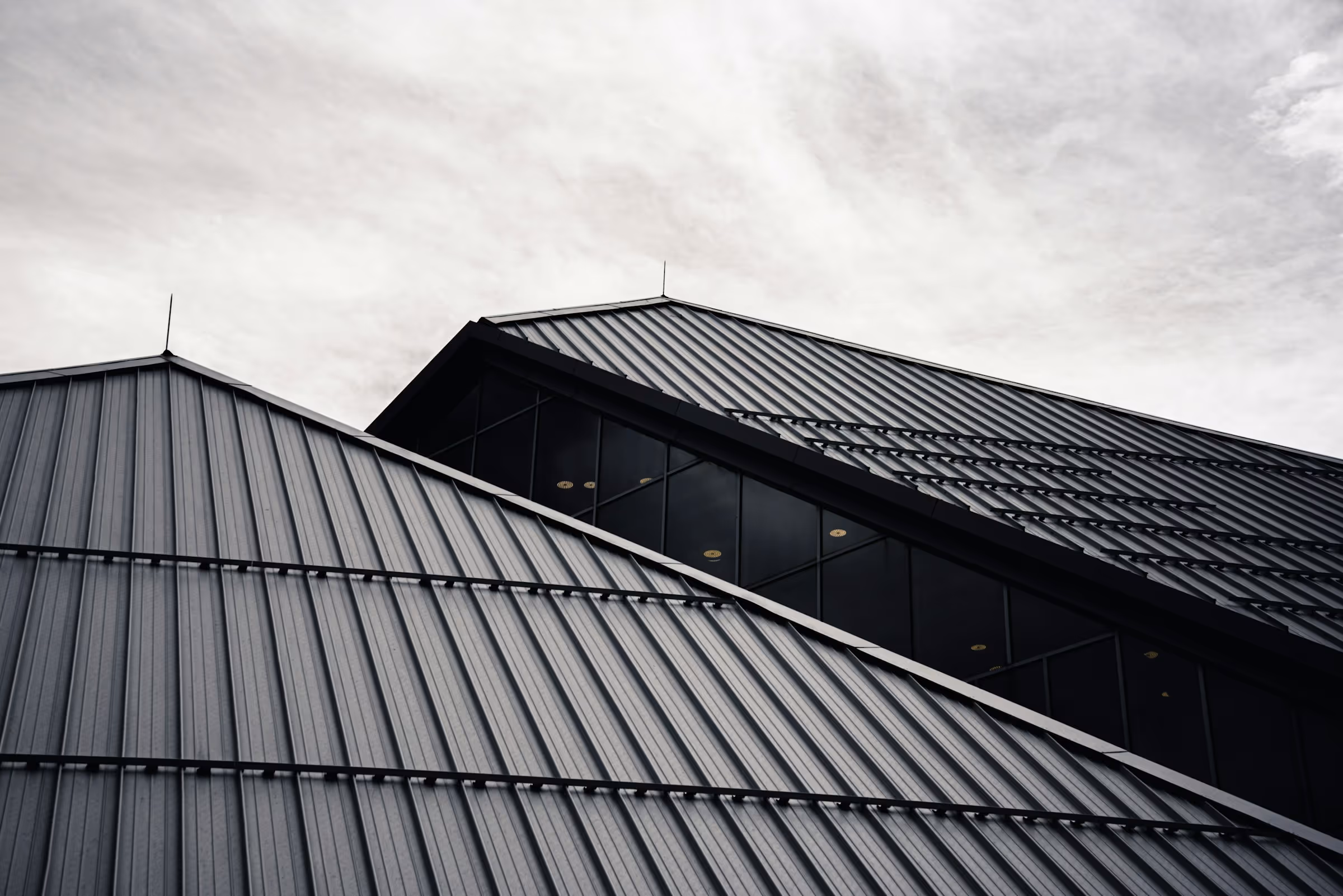 Modern building with sloped black metal roofs and large glass windows under a cloudy sky.