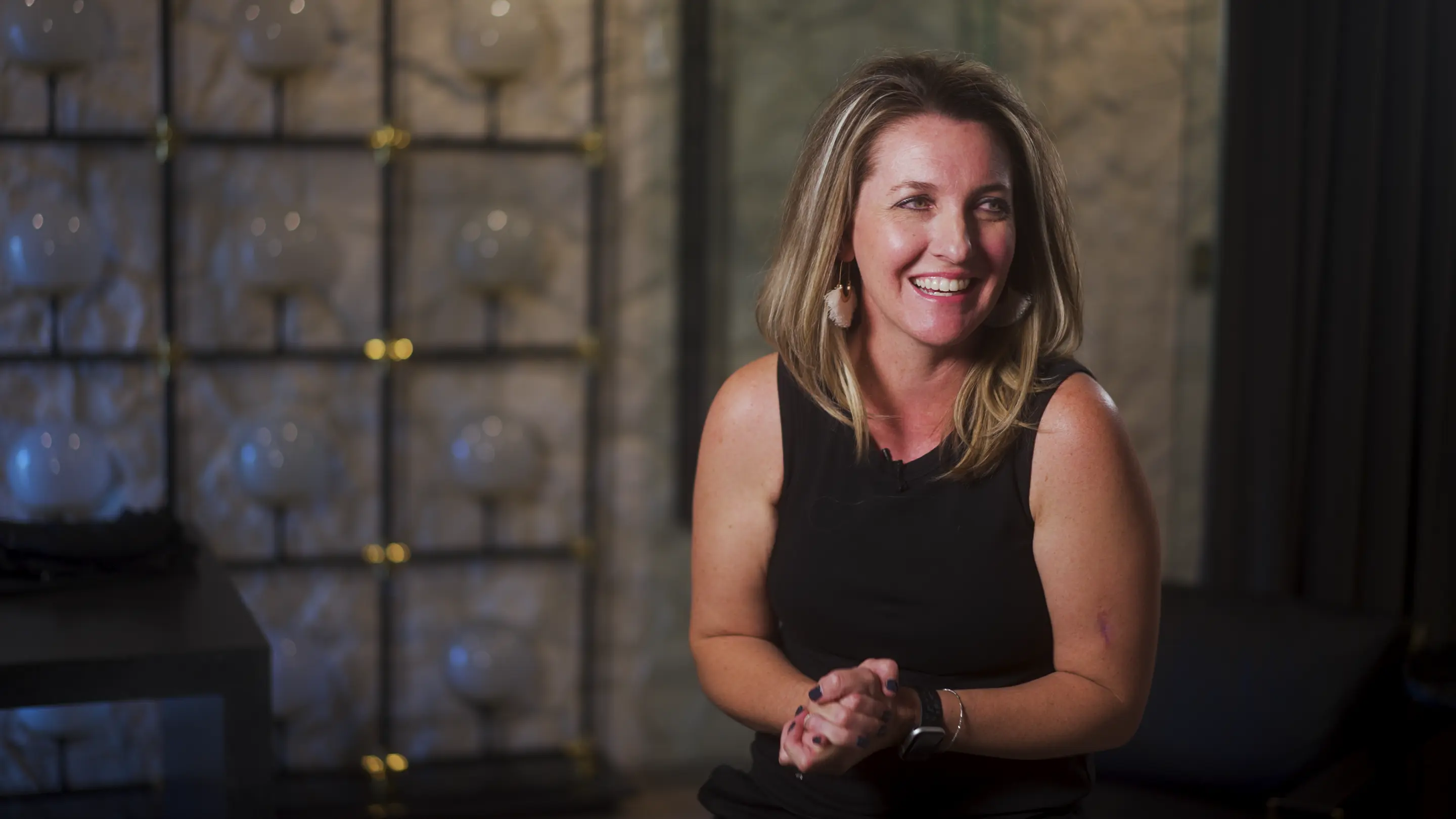 Smiling woman with blonde hair wearing a black sleeveless top and earrings, sitting indoors with a blurred background.