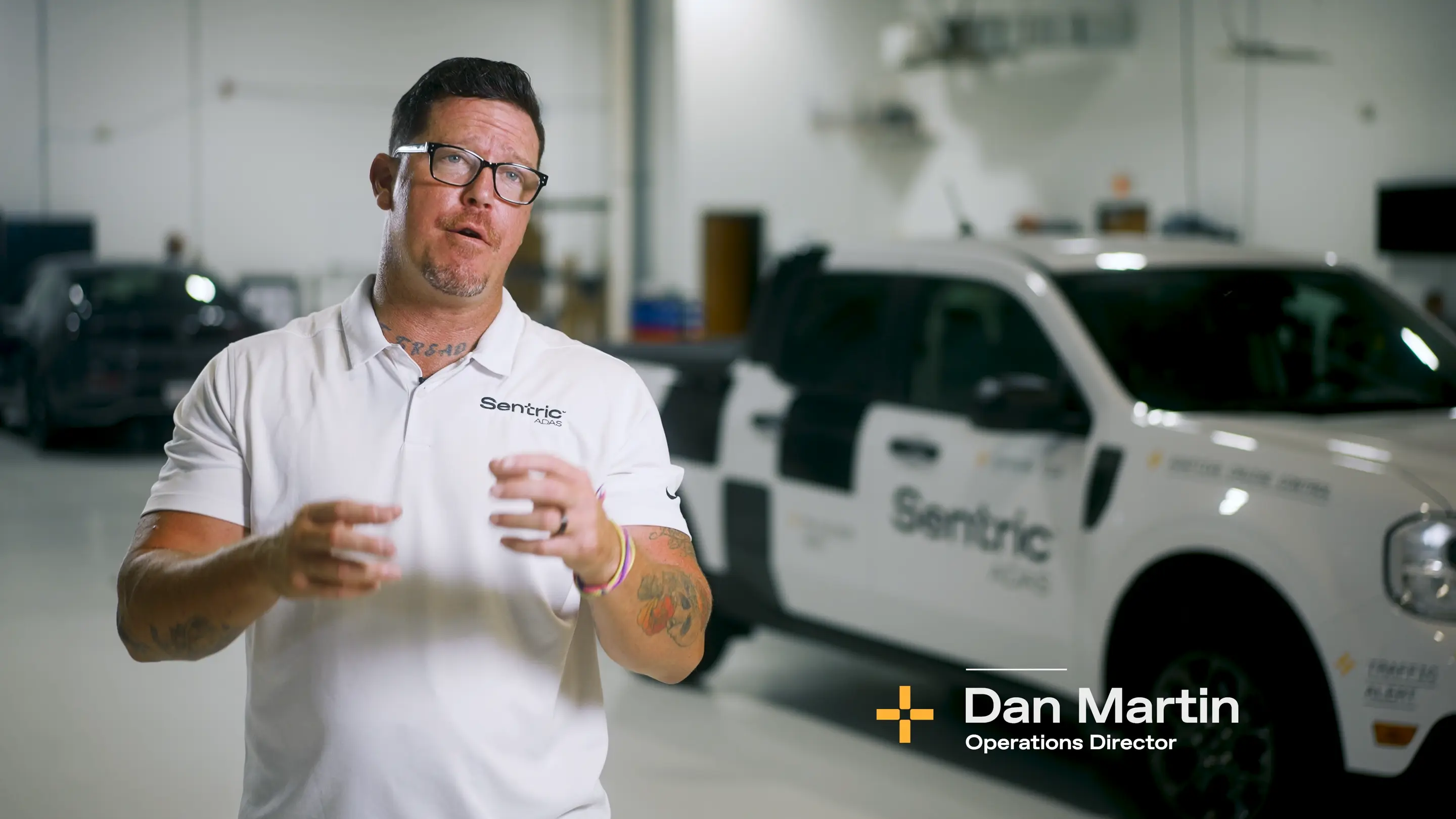 Man with glasses and tattoos wearing a white Sentric ADAS polo shirt speaking in a garage with a white Sentric vehicle in the background, labeled Dan Martin, Operations Director.