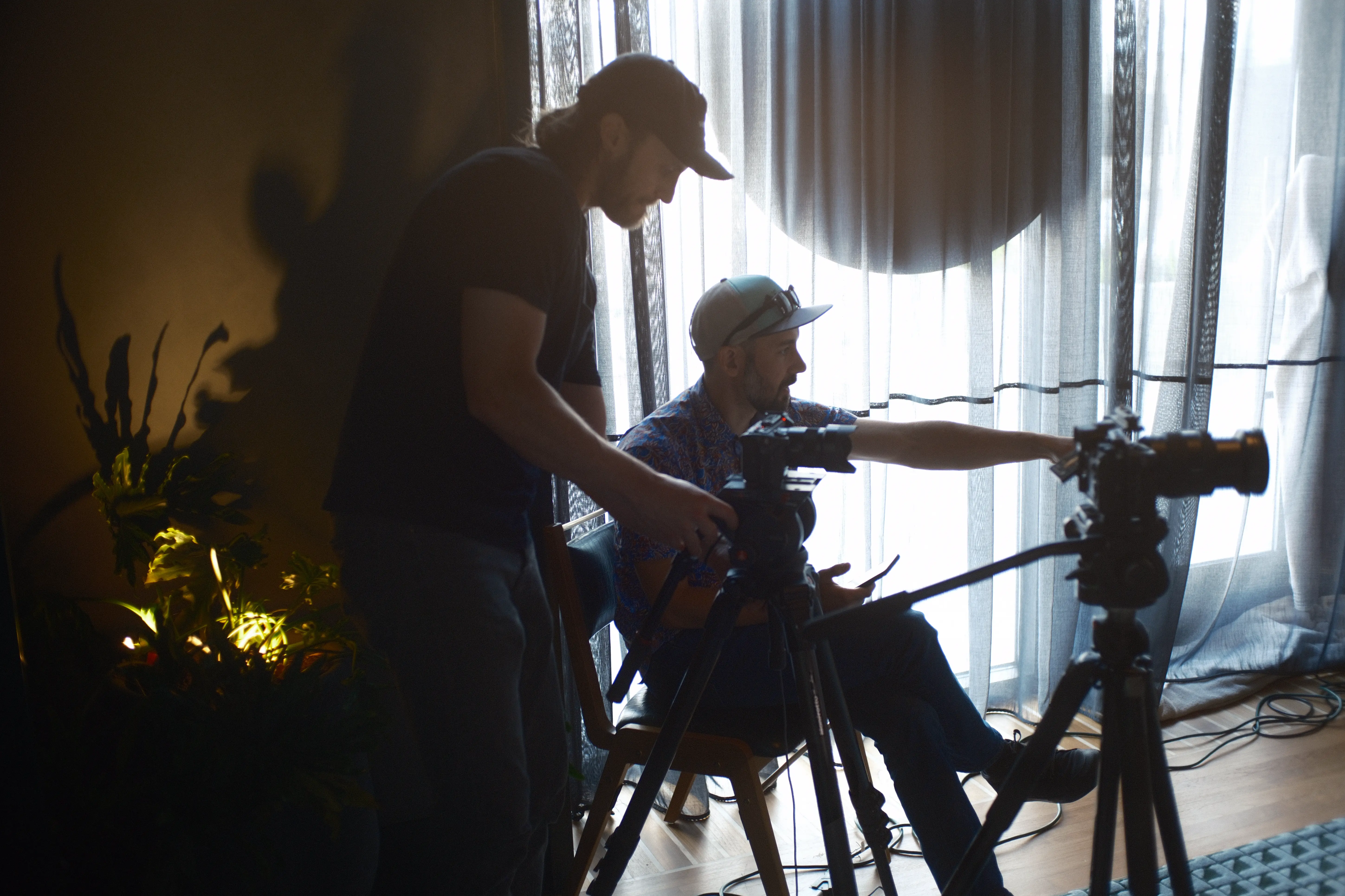 Two men operating cameras indoors near a window with sheer curtains, one seated pointing and the other adjusting equipment.