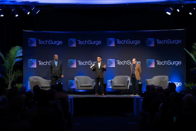 Three men standing on stage with TechSurge logos on backdrop, speaking to an audience.
