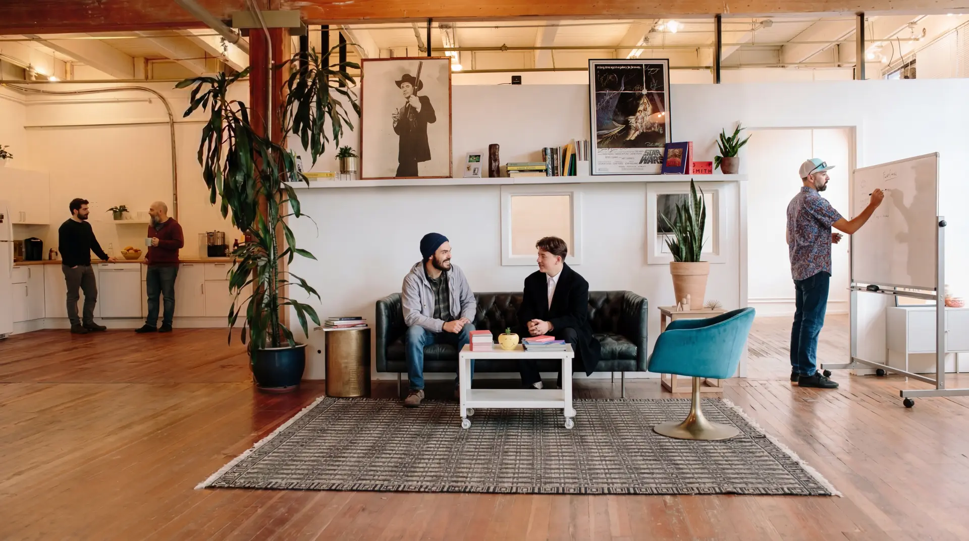Modern open office with wooden floors, two people chatting on a black couch, a man writing on a whiteboard, and two men talking in the kitchen area.