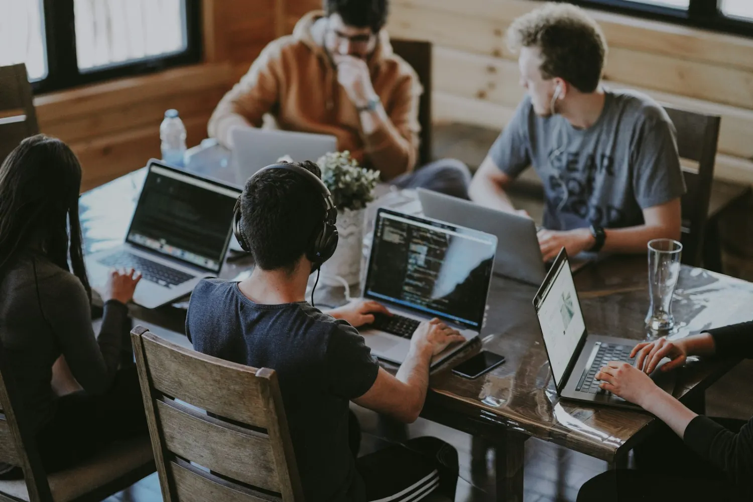 Group of young adults working on laptops around a wooden table in a casual indoor setting.
