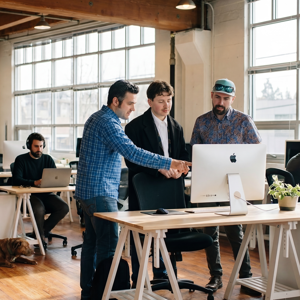 Three men collaborating around a desktop computer in a modern office with large windows, a man working on a laptop in the background, and a dog lying on the floor.