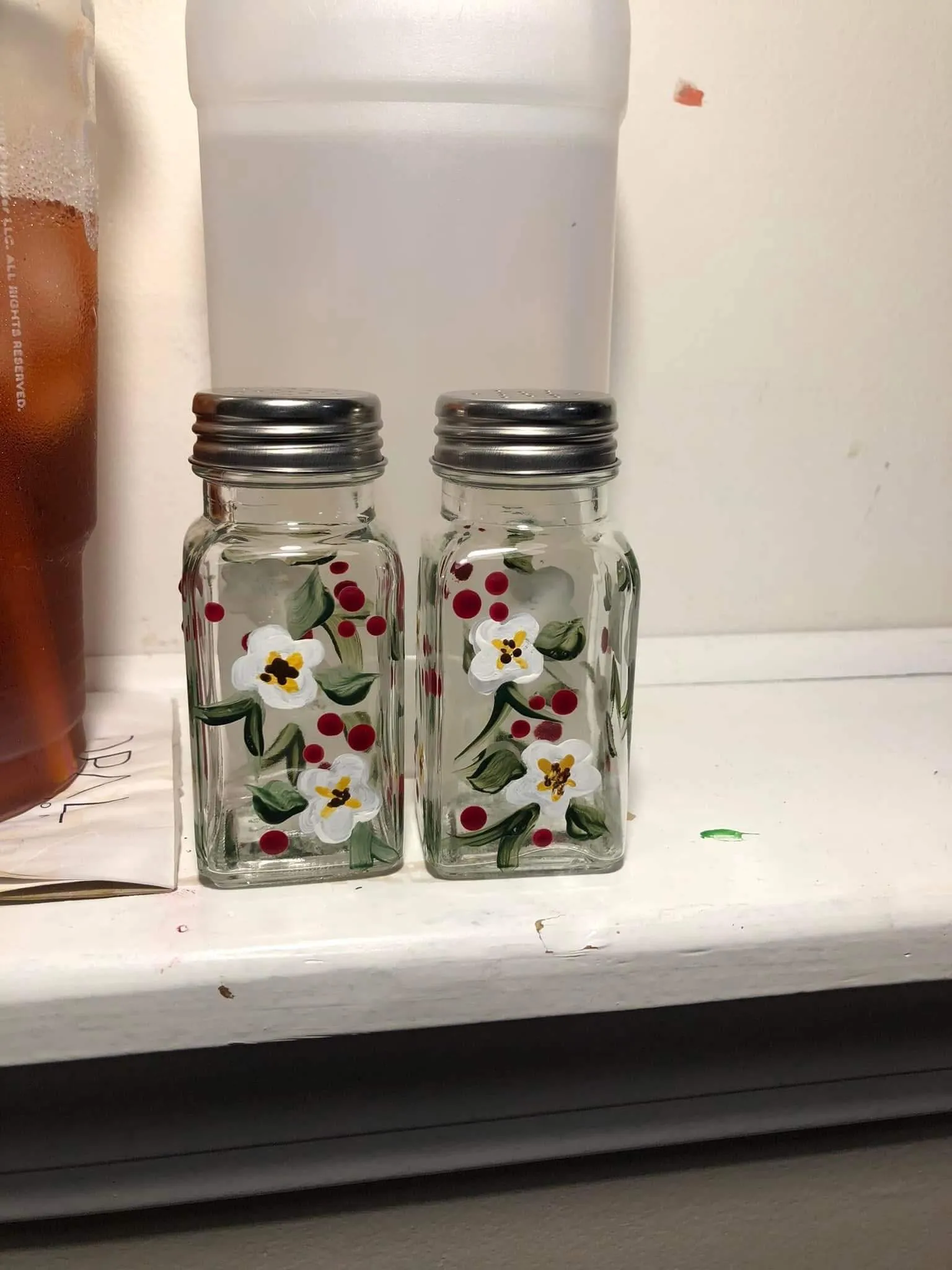Pair of clear glass salt and pepper shakers hand-painted with white flowers, green leaves, and red berries, placed on a windowsill.