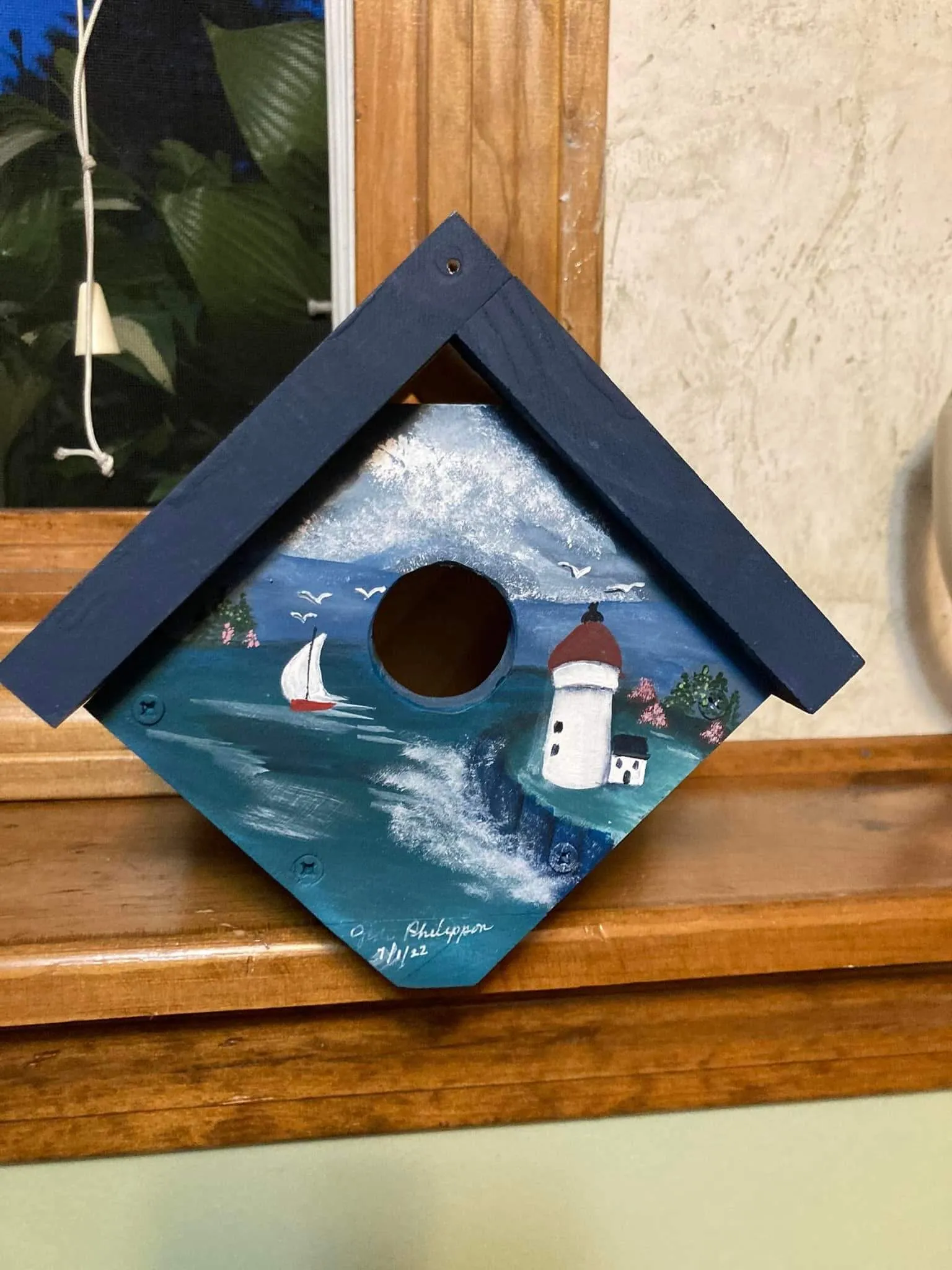 A blue painted wooden birdhouse with an ocean scene depicted on it of a lighthouse, waves, and a small sailboat.