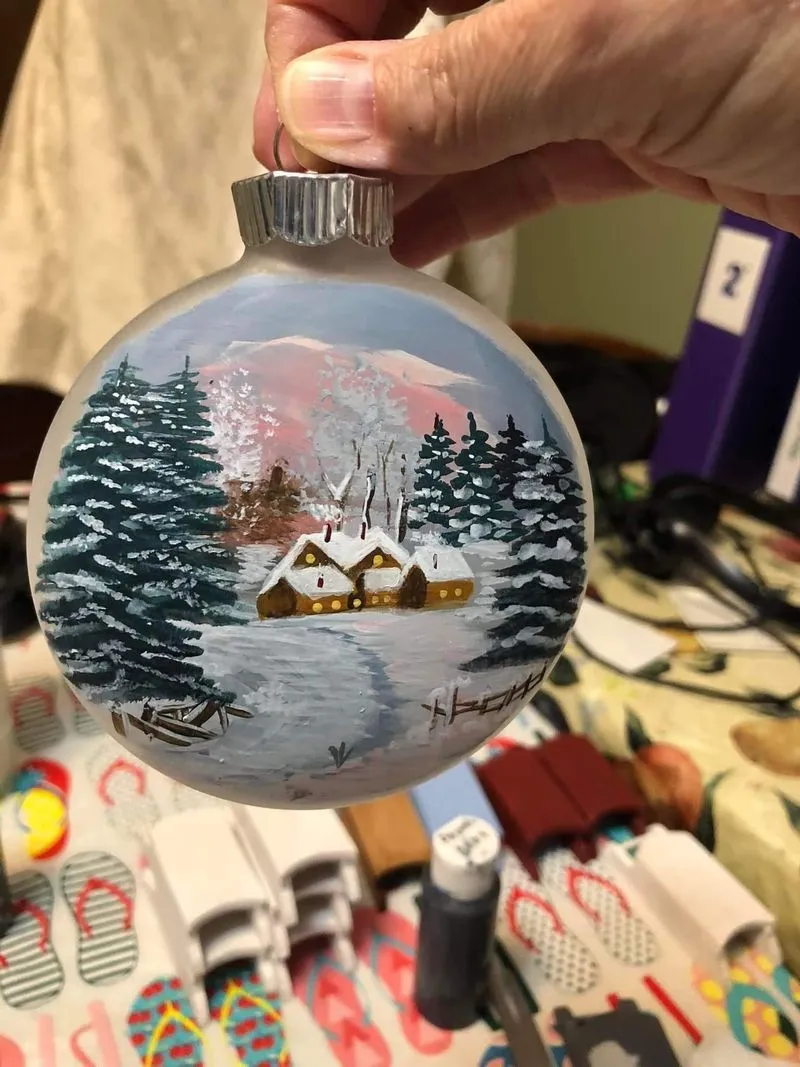 An ornament painted to look like a snowy winter scene with several houses, trees, and snow covering the ground.