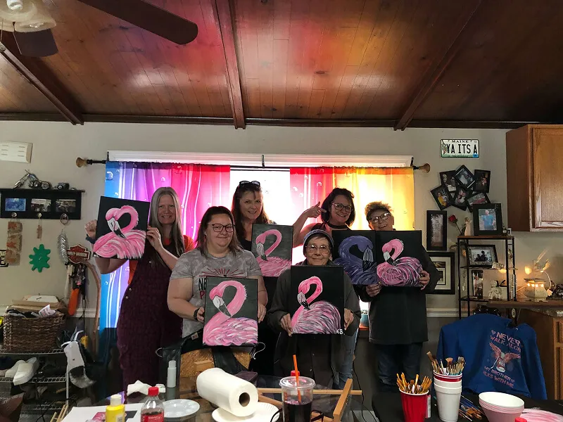 A group of six women each holding a painting of a flamingo on canvas. in front of them are paint brushes and easels to show that they painted these flamingos themselves.