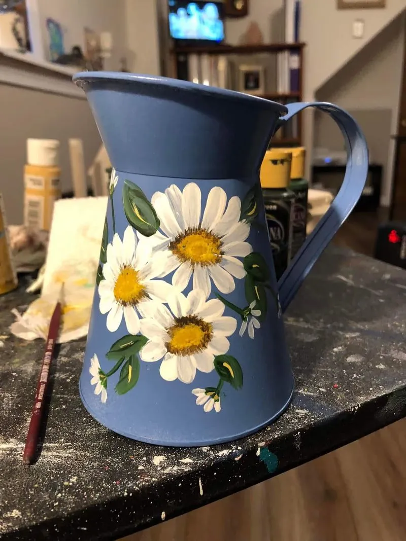 A light blue metal pitcher hand-painted with large white daisies and green leaves, resting on a speckled tabletop.