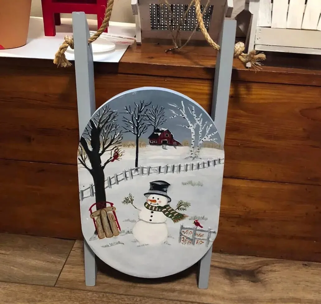 A wooden sled painted in hues of light gray and blue that has a winter scene on it of a snowman in the snow next to a leafless tree.