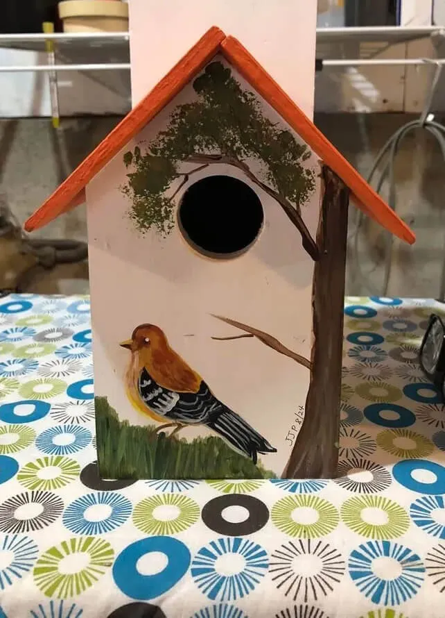 A wooden birdhouse with an orange roof. Painted on the front is a bird sitting on the grass beneath a tree.