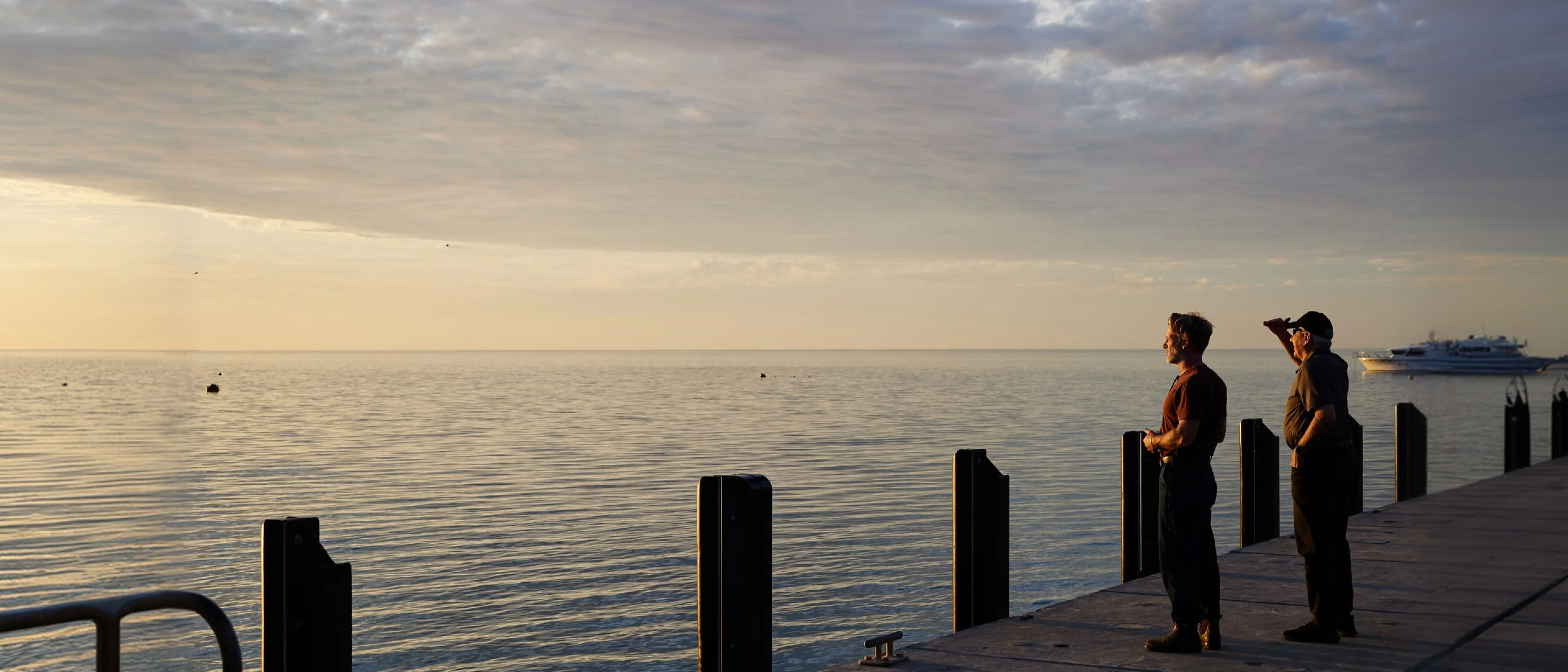 Two people standing on a pier overlooking a calm ocean with a distant boat during sunset.