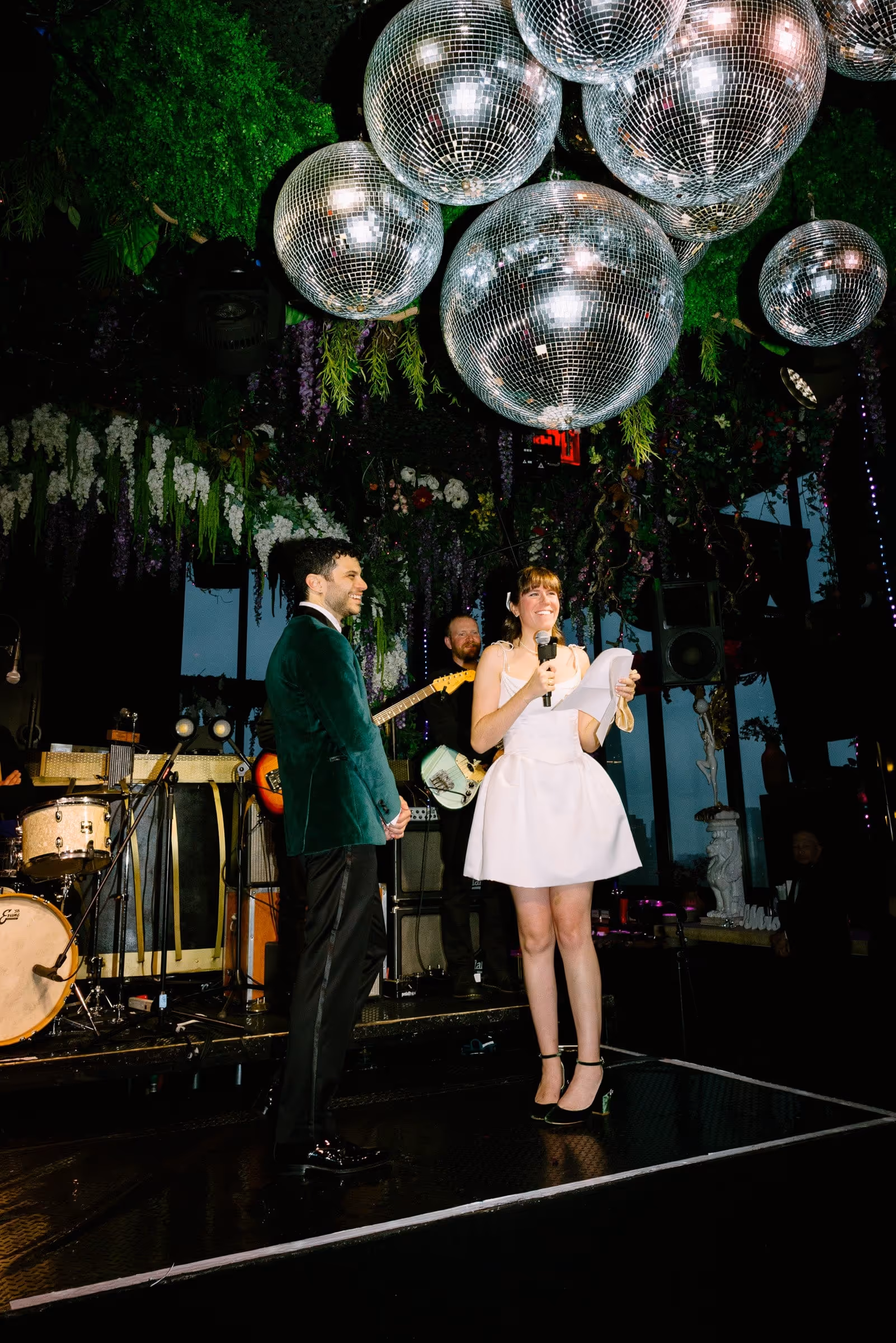 A bride in a short white dress holding a microphone and paper, standing next to a groom in a green velvet jacket under large disco balls at a music stage with a band.