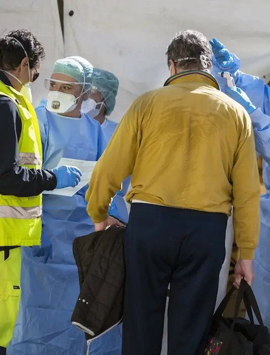 A checkpoint to control the spread of a pandemic, with three figures in extensive PPE checking the vitals of two people seeking entry