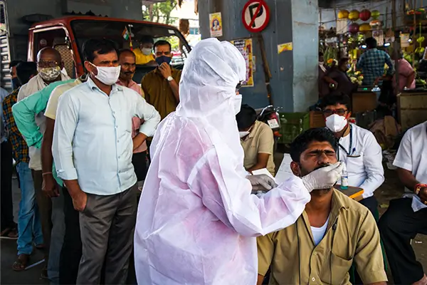 A healthcare worker dressed in full, disposable PPE tests an Indian man during the pandemic.  A long line of men crowd closely together waiting to be tested for COVID during the pandemic.
