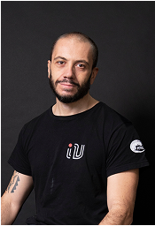 A man with a beard and a shaved head, wearing a black t-shirt, posed against a dark background. He is looking directly at the camera.