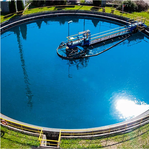 An aerial view of a water treatment facility featuring a circular, turquoise water tank with a walkway and equipment in the center, surrounded by green grass and trees.