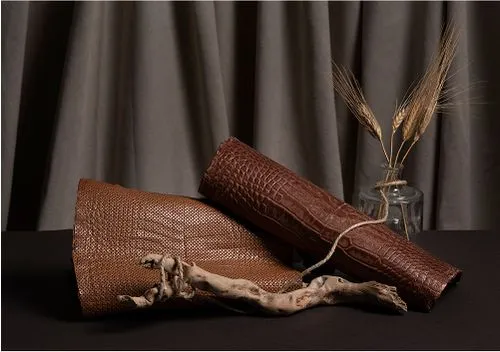 Two textured brown leather rolls and a piece of driftwood arranged on a dark surface, with a glass vase containing dried wheat in the background, against a backdrop of gray fabric.