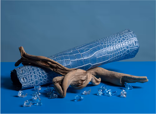 A blue crocodile-embossed leather roll placed on a blue surface, accompanied by a piece of driftwood and scattered clear plastic pieces.