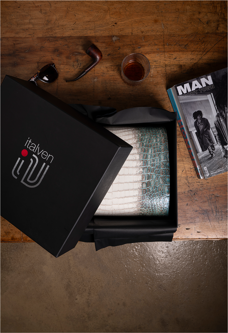 A black box with the logo 'italven' partially opened to reveal a textured item wrapped in black tissue paper. Nearby, there are sunglasses, a tobacco pipe, a glass of amber liquid, and a magazine titled 'MAN' on a wooden surface.