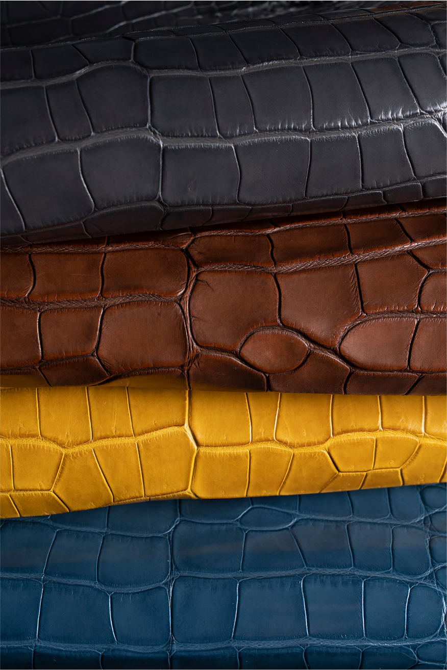 Four sheets of leather in different colors: dark gray, brown, mustard yellow, and dark blue, each featuring a textured crocodile pattern, stacked neatly on top of one another.