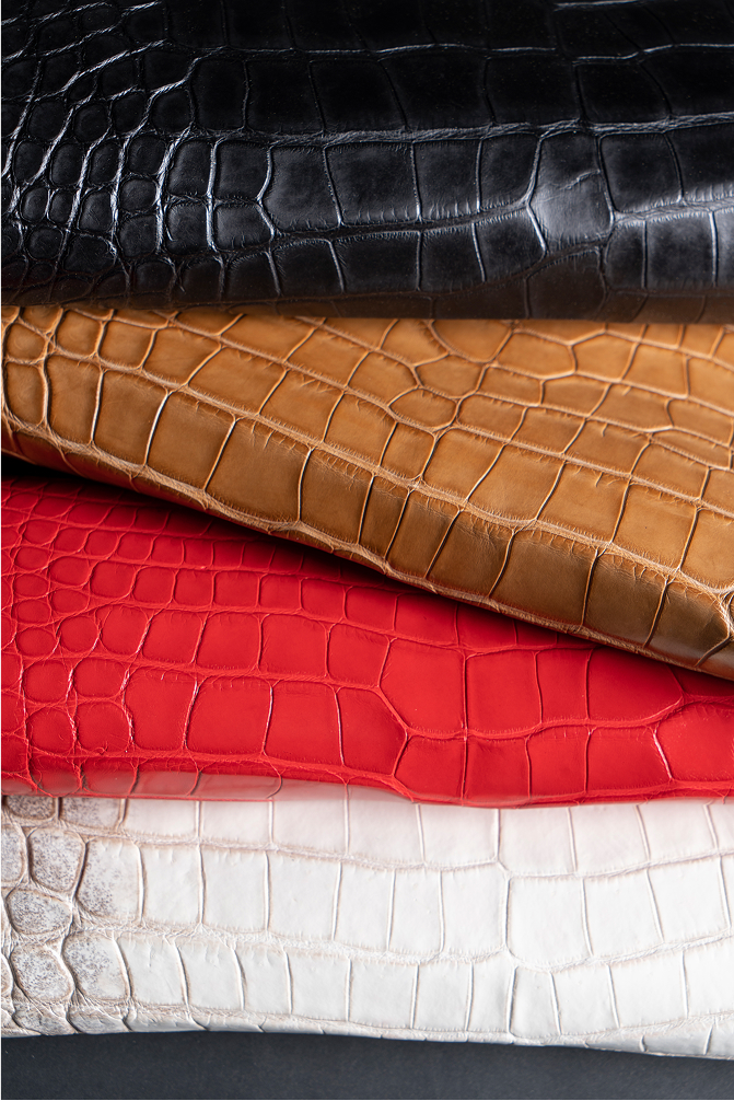 Four pieces of textured leather stacked. The top layer is black, followed by a light brown, red, and finally a white layer. Each piece showcases a distinct crocodile pattern.