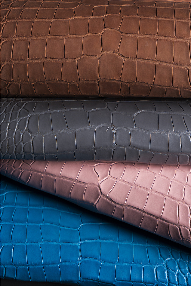 Four pieces of textured fabric in different colors: brown, gray, pink, and blue, all resembling crocodile skin. The fabrics are stacked on top of each other, showcasing their unique patterns.