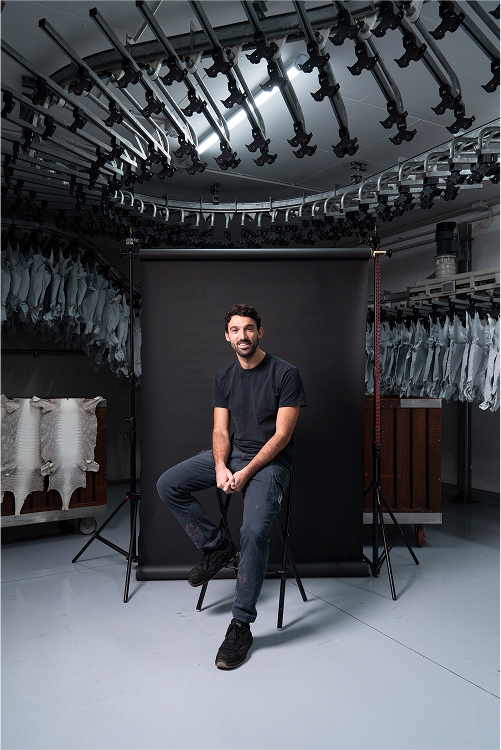 A man sitting on a stool in front of a black backdrop inside a facility with a circular arrangement of equipment above. White materials are visible in the background, hinting at an industrial setting.