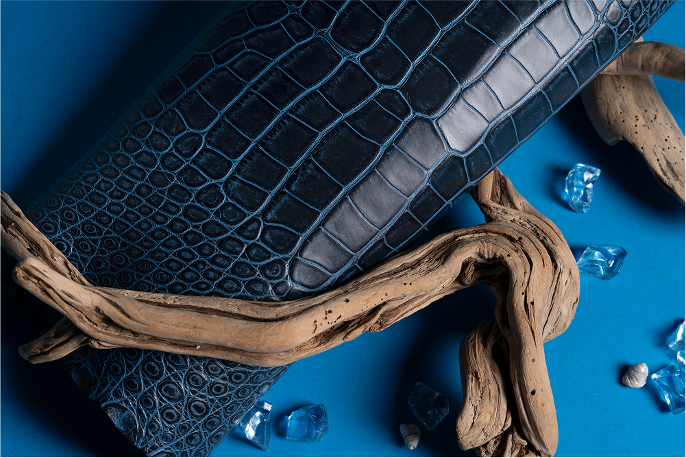 A rolled piece of blue crocodile-embossed leather placed on a blue background, surrounded by driftwood and decorative glass stones.