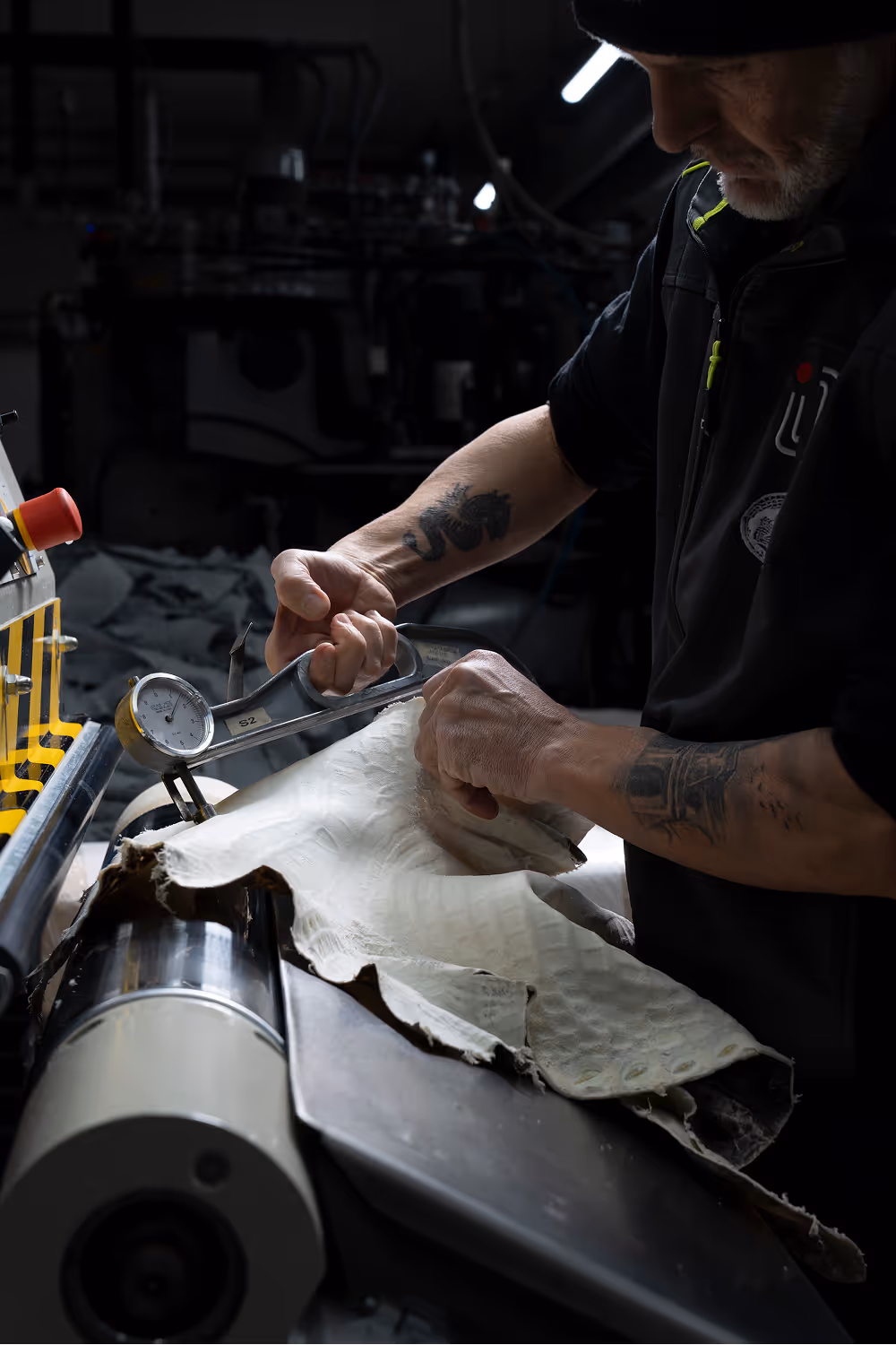 A worker measuring a piece of leather using a caliper, surrounded by rolls of leather in a workshop. The focus is on the precision of the measurement, and the workspace is dimly lit.