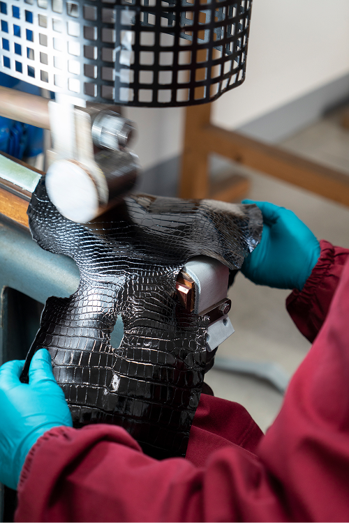 A person wearing blue gloves is working with a piece of black crocodile-patterned leather on a machine. The setting appears to be a workshop, with tools and materials in the background.