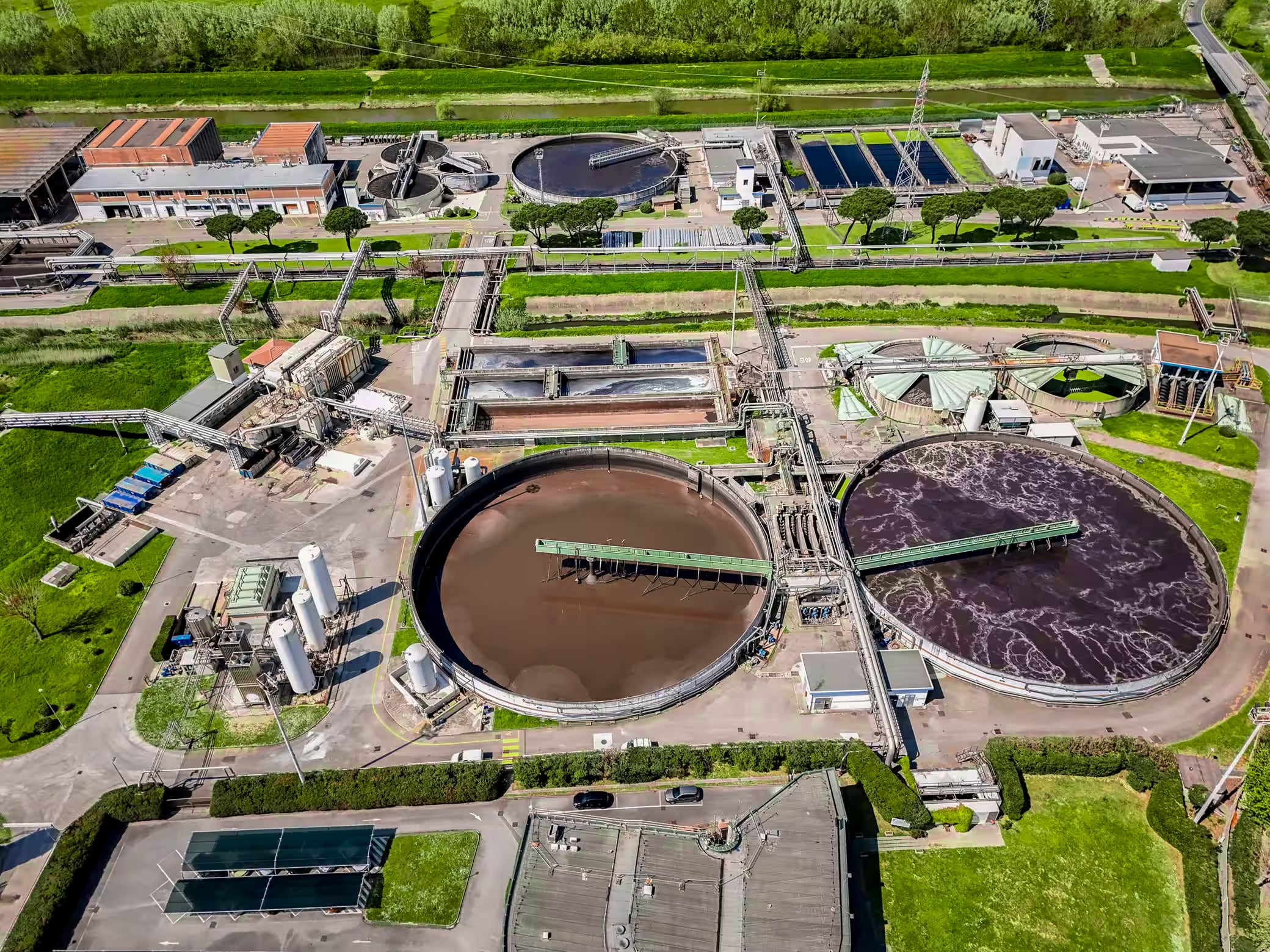 An aerial view of a wastewater treatment facility featuring several large circular tanks and various industrial structures, surrounded by green grass and trees.