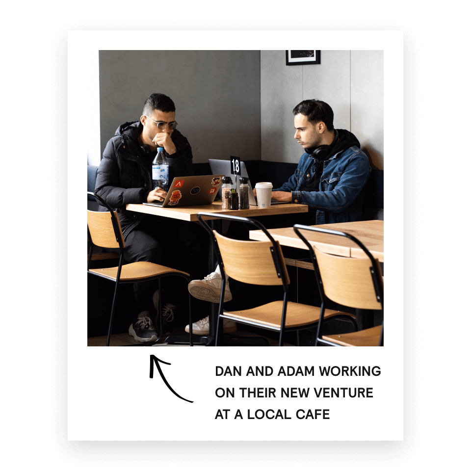 Two men sitting at a cafe table, working on laptops with drinks and condiments on the table.