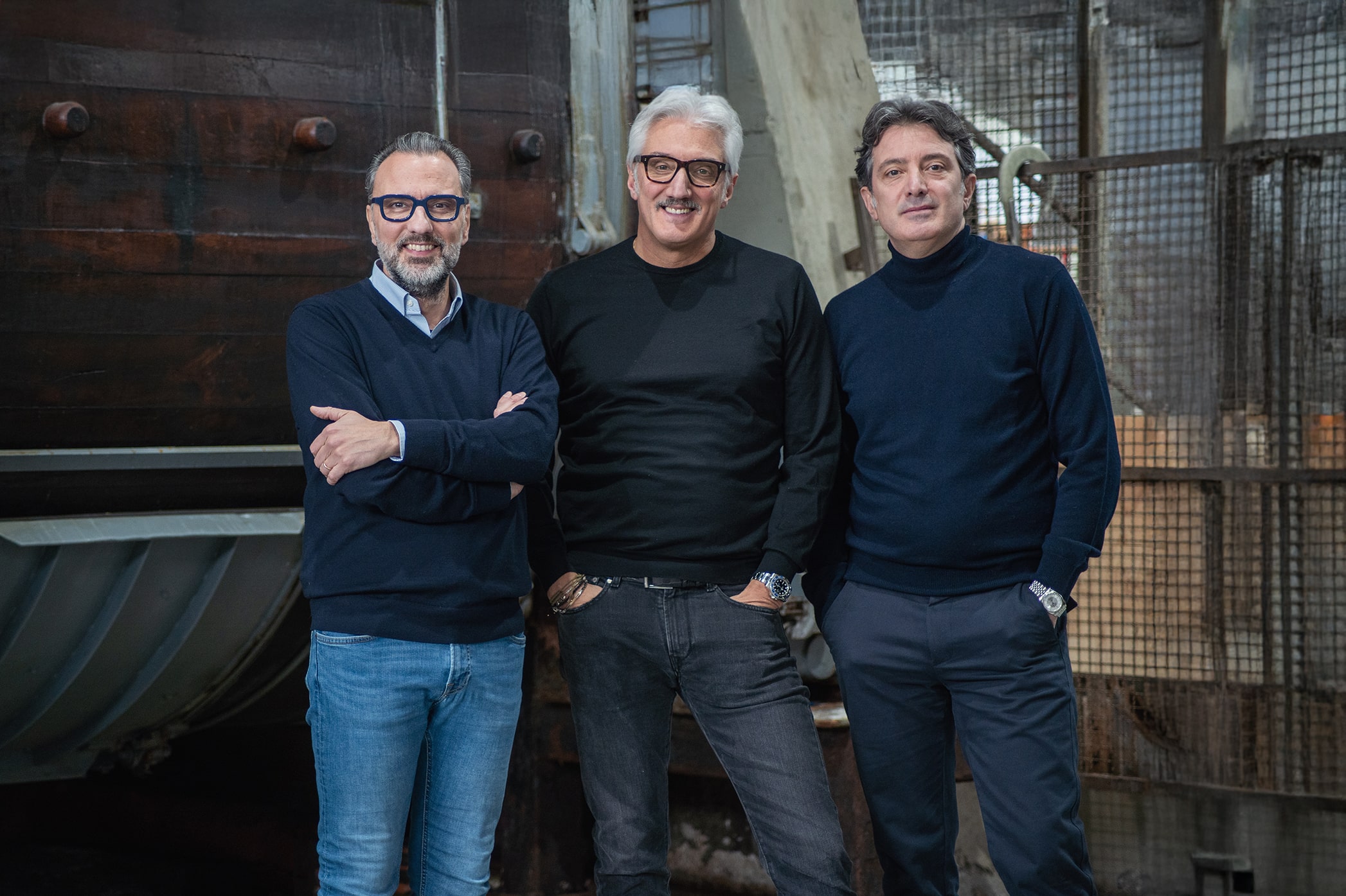 Three men standing indoors against industrial background, smiling and dressed in dark sweaters and casual pants.