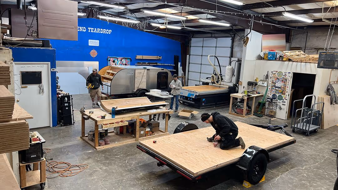 The interior of a large workshop where a person is working on a project next to a vehicle frame and workbenches.