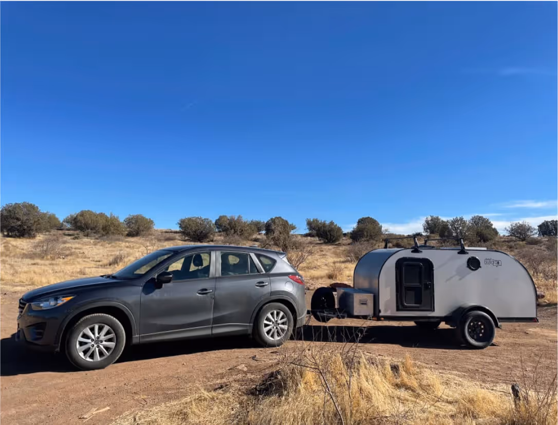 A pickup truck towing a silver camping trailer is parked on a dirt road in a sunny, arid landscape.