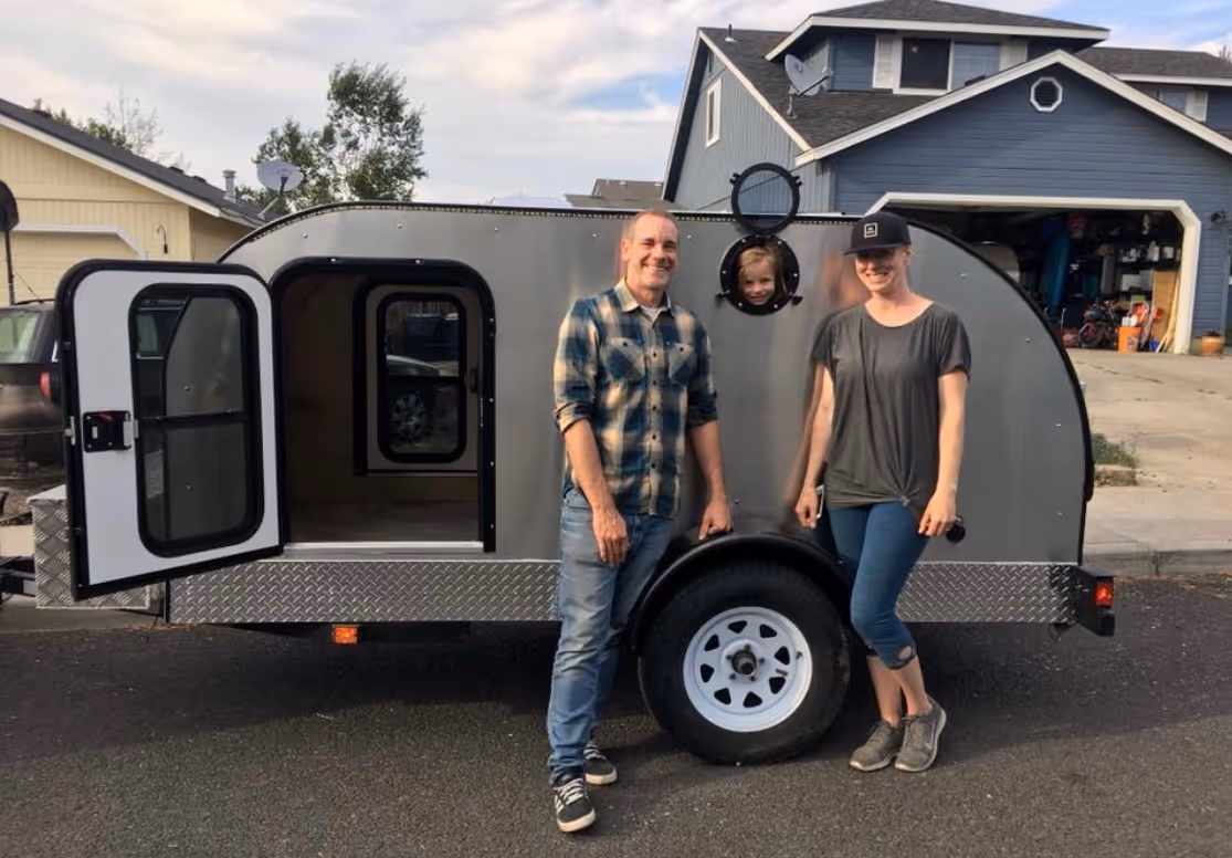 A man stands proudly next to his camping trailer, which is hitched to a truck.