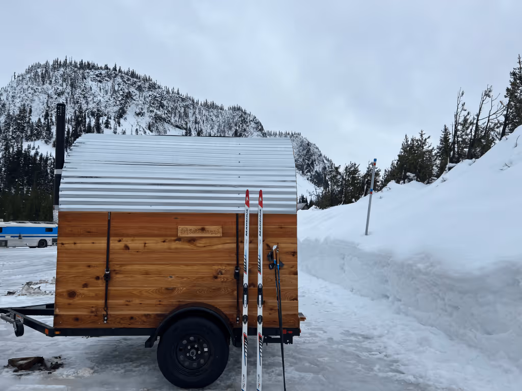 A mobile sauna on a trailer equipped with skis instead of wheels, parked in a deep snowy field.