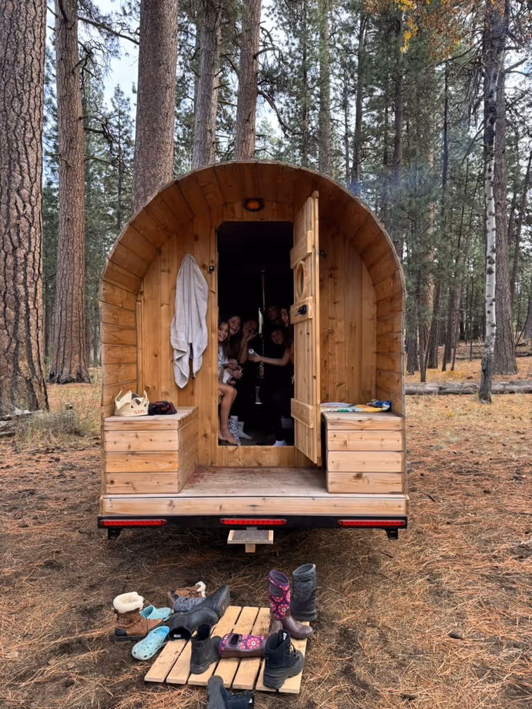 A mobile barrel sauna on a trailer is adorned with festive string lights in a wooded setting.