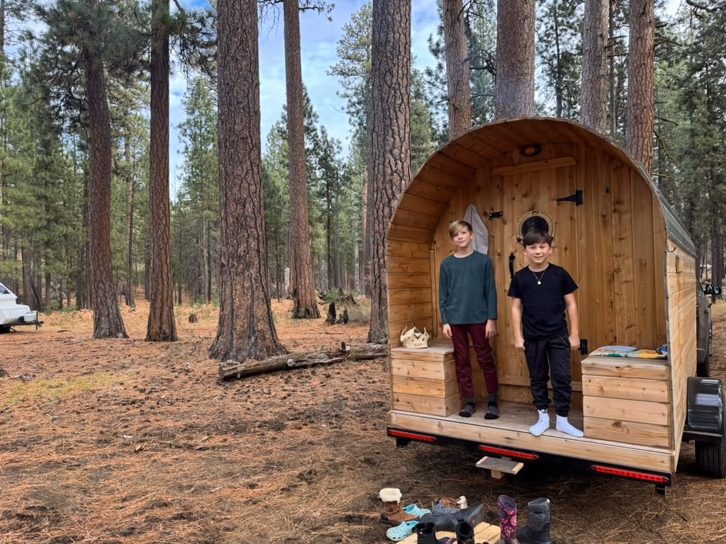 A barrel sauna is nestled among tall pine trees, with two people standing in its circular doorway.