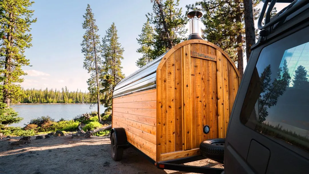 A barrel sauna sits on a deck overlooking a calm lake, surrounded by a forest and mountains.