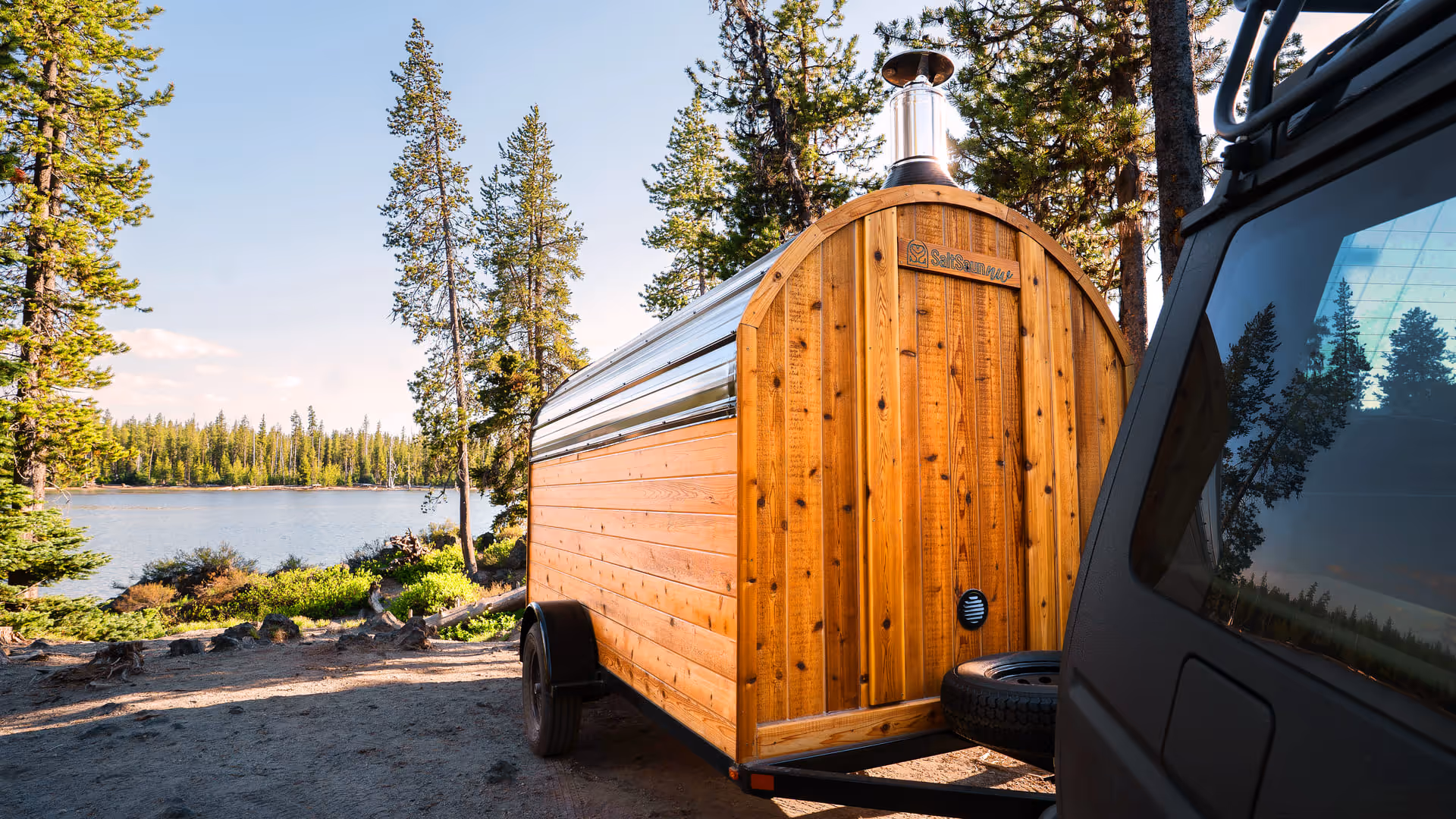A barrel sauna sits on a deck overlooking a calm lake, surrounded by a forest and mountains.