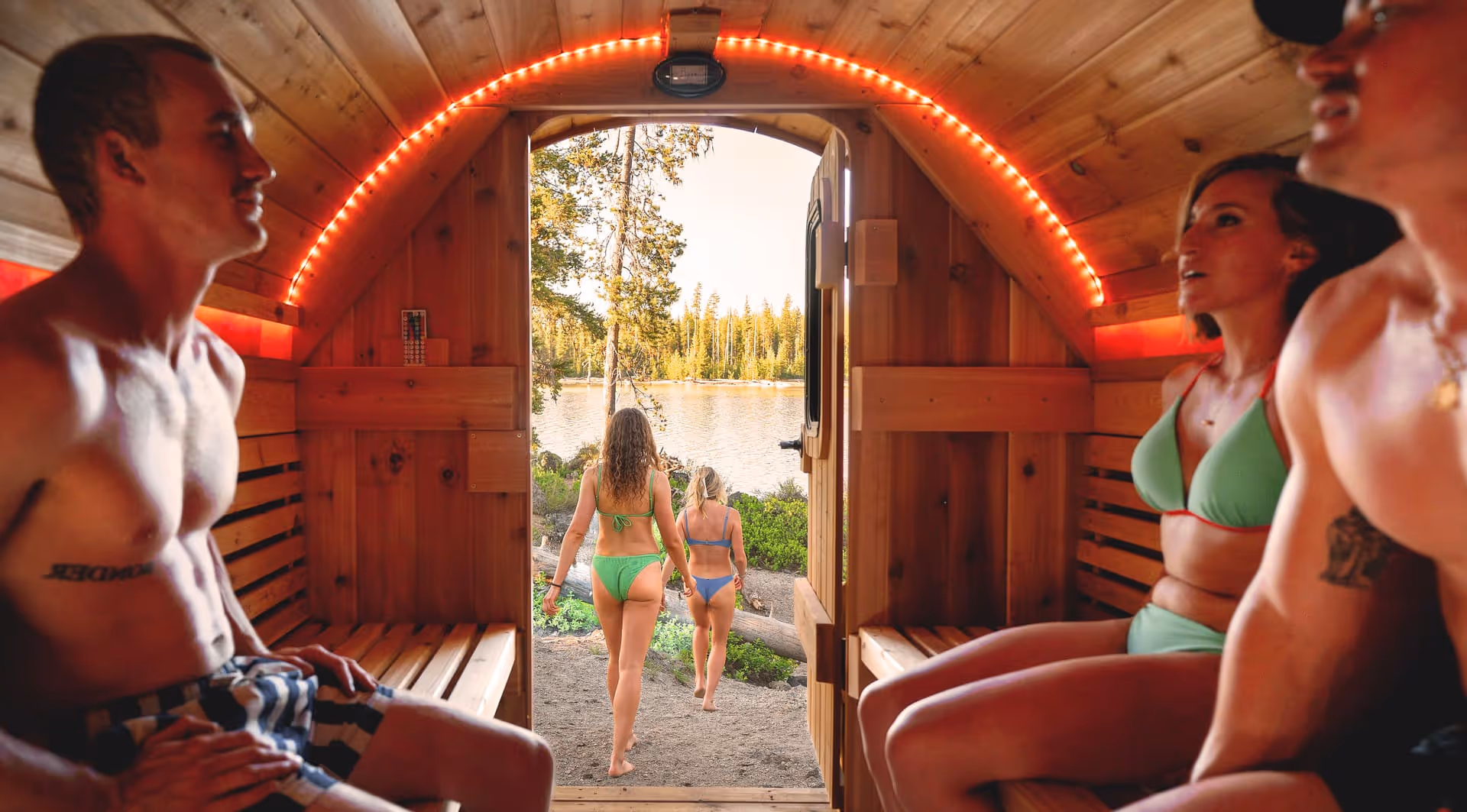 A group of people in swimsuits relaxes inside a sauna, seen from the back looking out the open door to a forest.