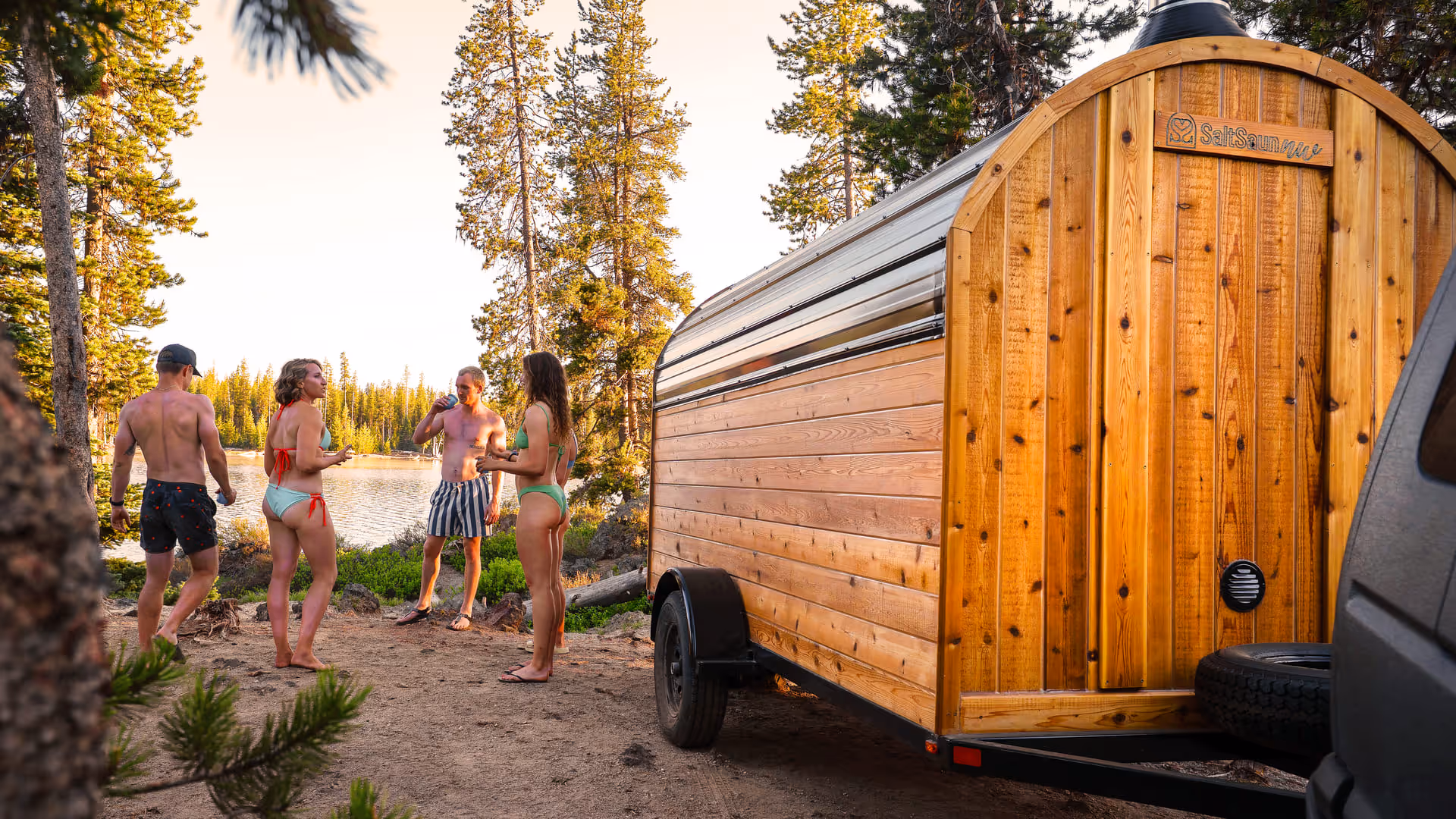 A side view of a mobile barrel sauna with a small porch, parked in a rustic, outdoor setting.