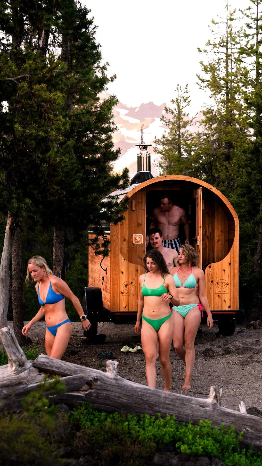 Group of people in swimsuits exiting a barrel-shaped outdoor sauna surrounded by pine trees, with snow-capped mountains in the background