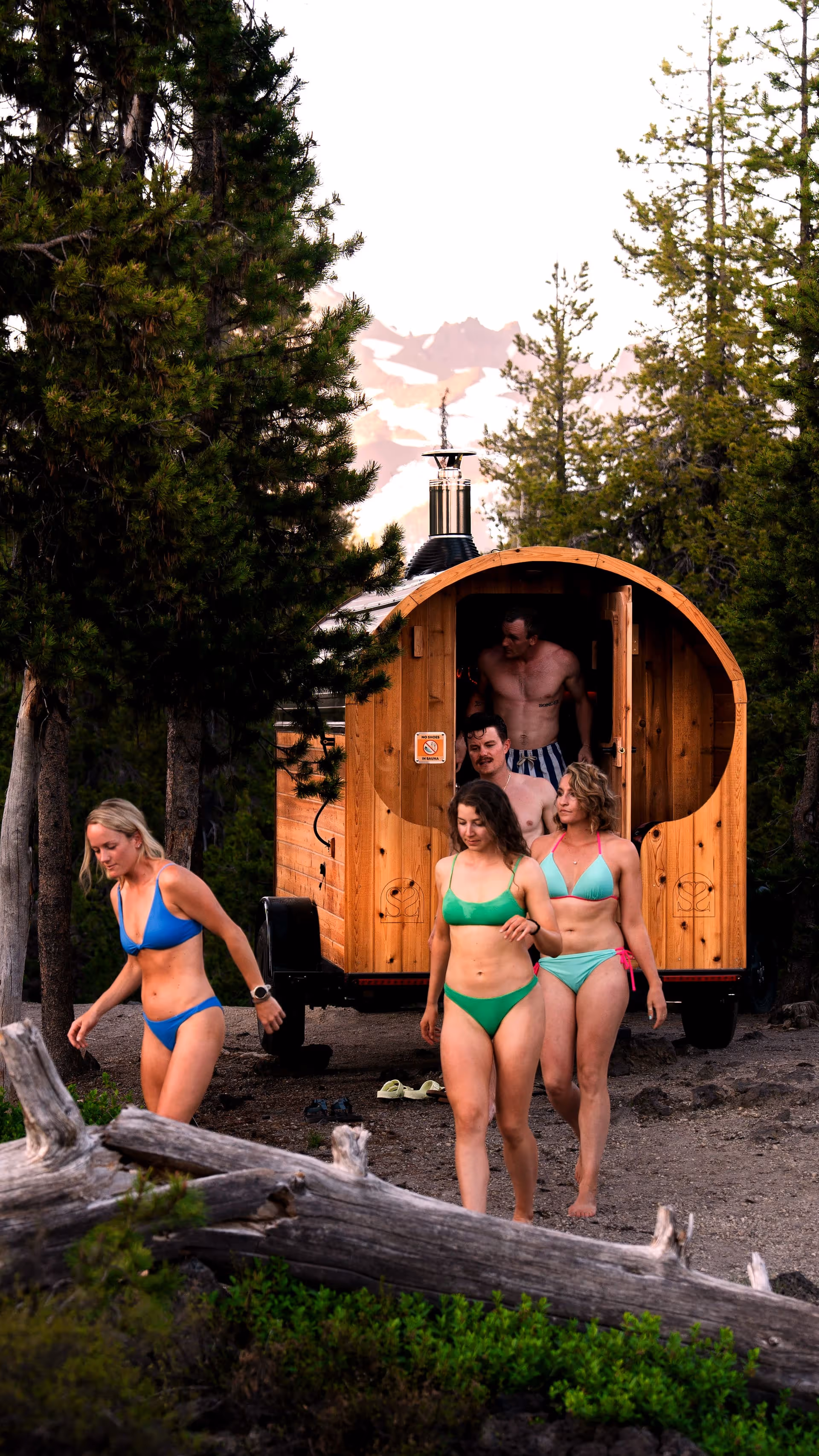 Group of people in swimsuits exiting a barrel-shaped outdoor sauna surrounded by pine trees, with snow-capped mountains in the background