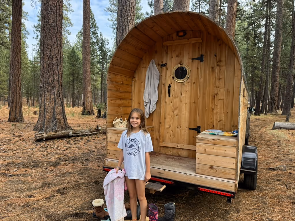 A woman and a small child sit on the steps of a mobile barrel sauna that is parked in a wooded area.