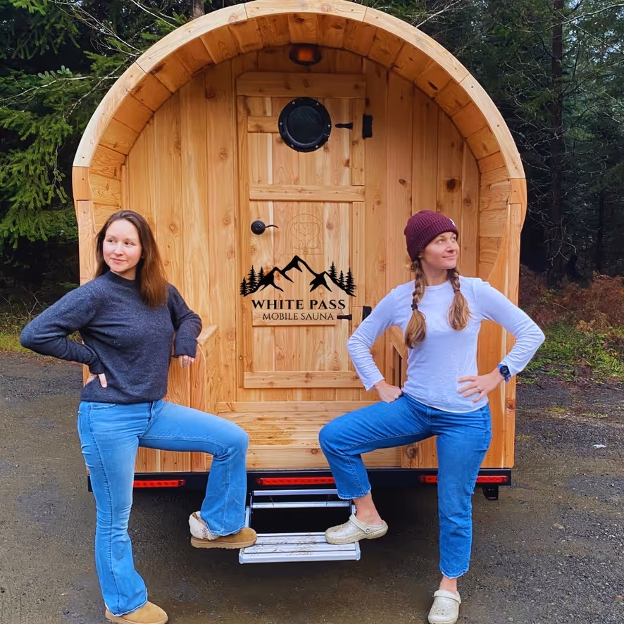 Two smiling women wearing beanies sit casually in the large, round doorway of a wooden barrel sauna.
