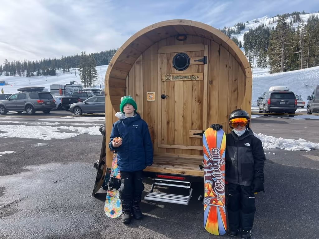 A group of friends in winter clothes poses happily in front of a mobile barrel sauna in a snowy parking lot.